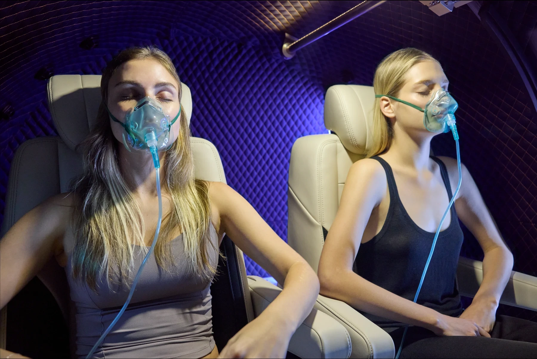 Two guests reclined side by side in a chamber cabin, oxygen masks on